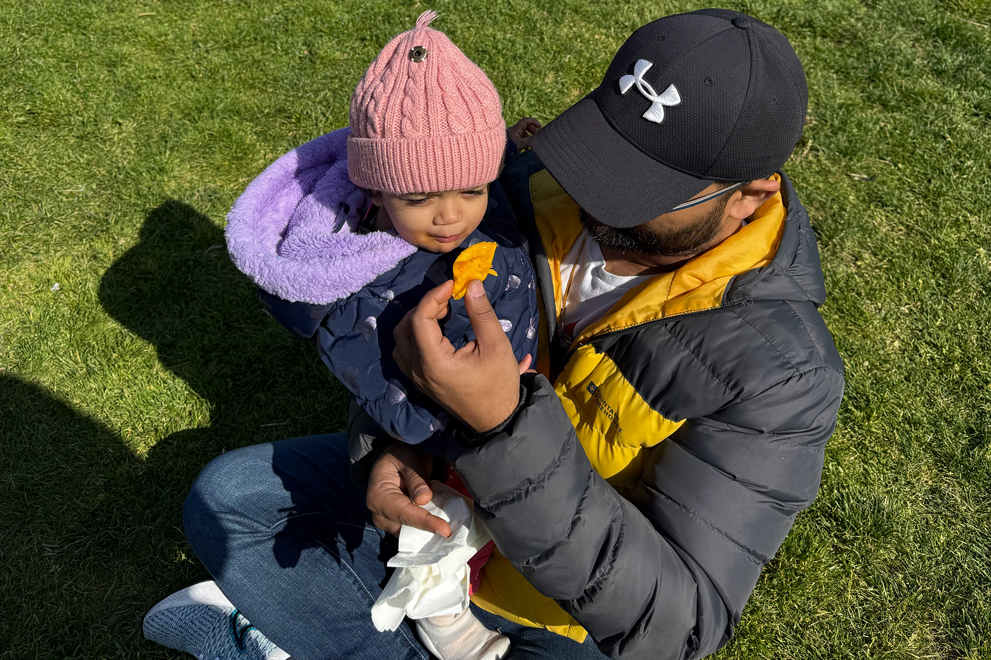 A father holding a young toddler feeds the child a piece of mango.