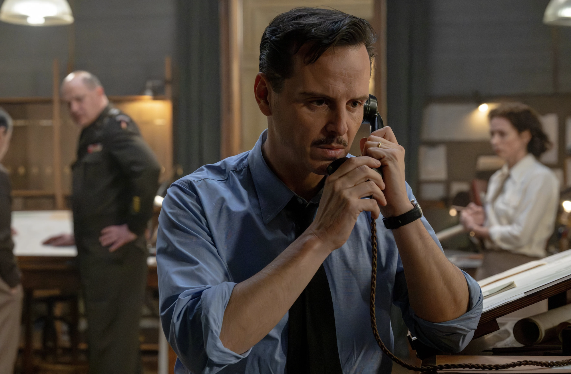A middle-aged white man with a mustaches, wearing 1940s-era blue shirt and tie, clutches a phone receiver to his left ear. A man in military uniform and a woman wearing a white blouse can be seen out of focus behind him.