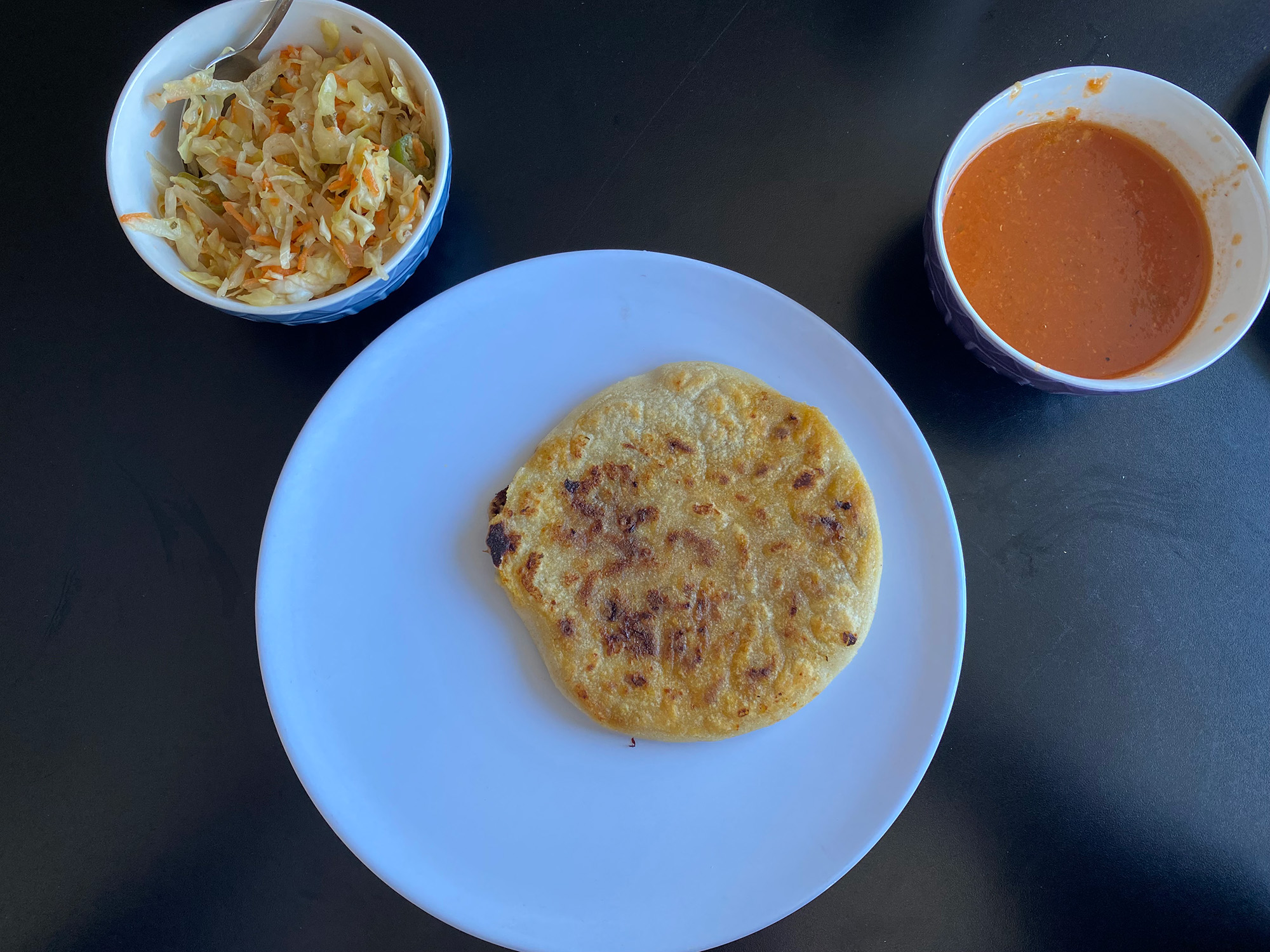 A pupusa on a white plate, with a small tub of curtido and a bowl of salsa on the side.