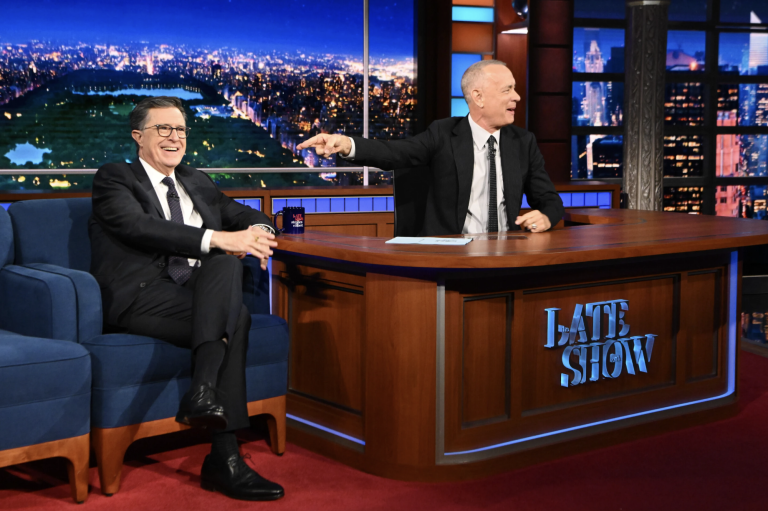 Two white men in suits sit near a large desk labeled LATE SHOW in a TV studio.