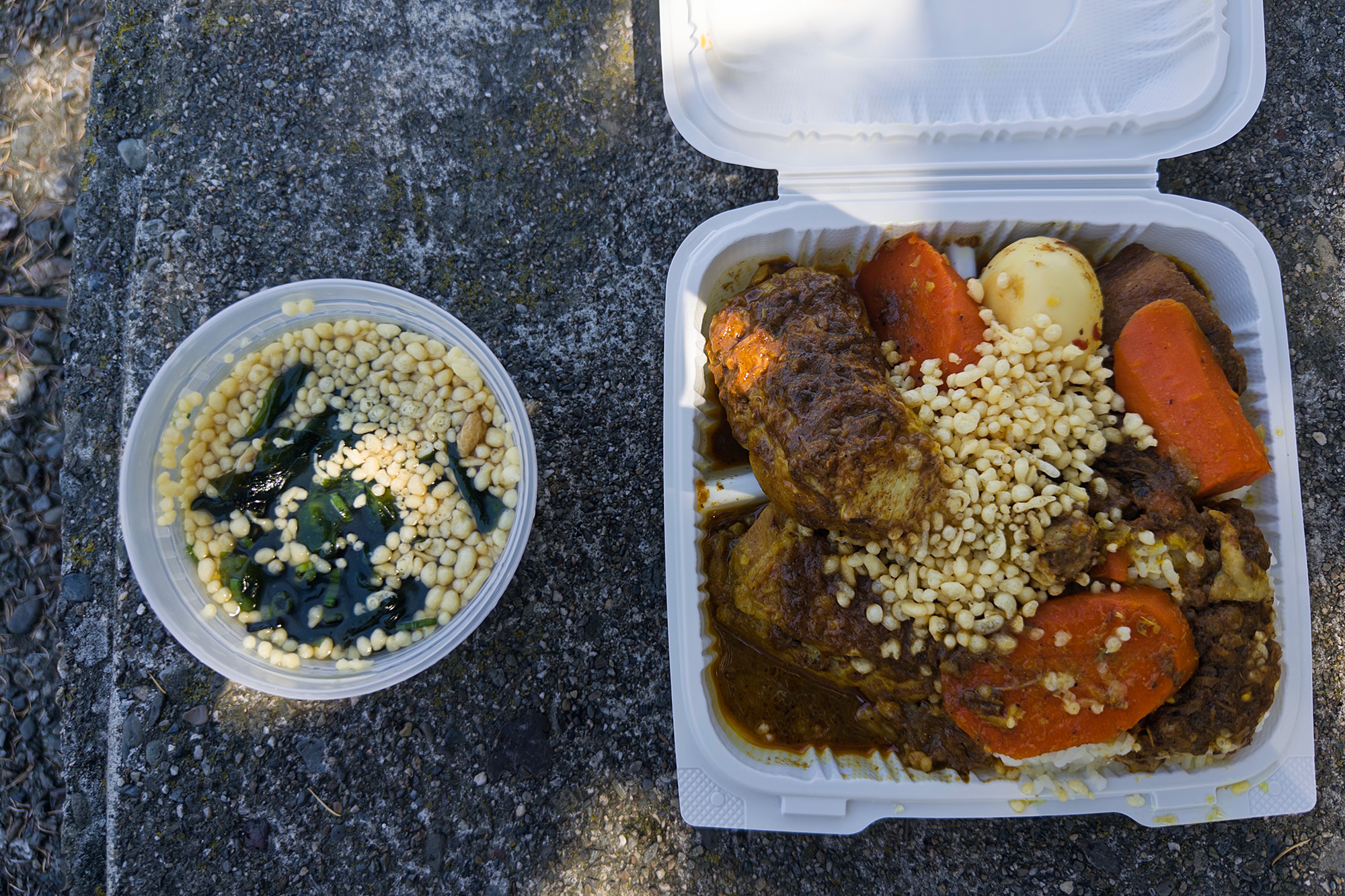 Takeout containers of soba and curry chicken against a concrete backdrop.