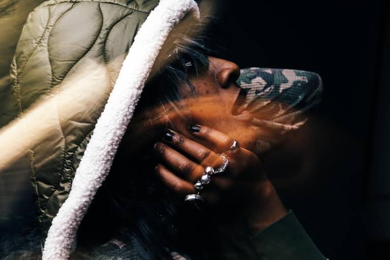 A Black face framed by a hand with multiple rings and a puffy hoddie, looking upward and to the right