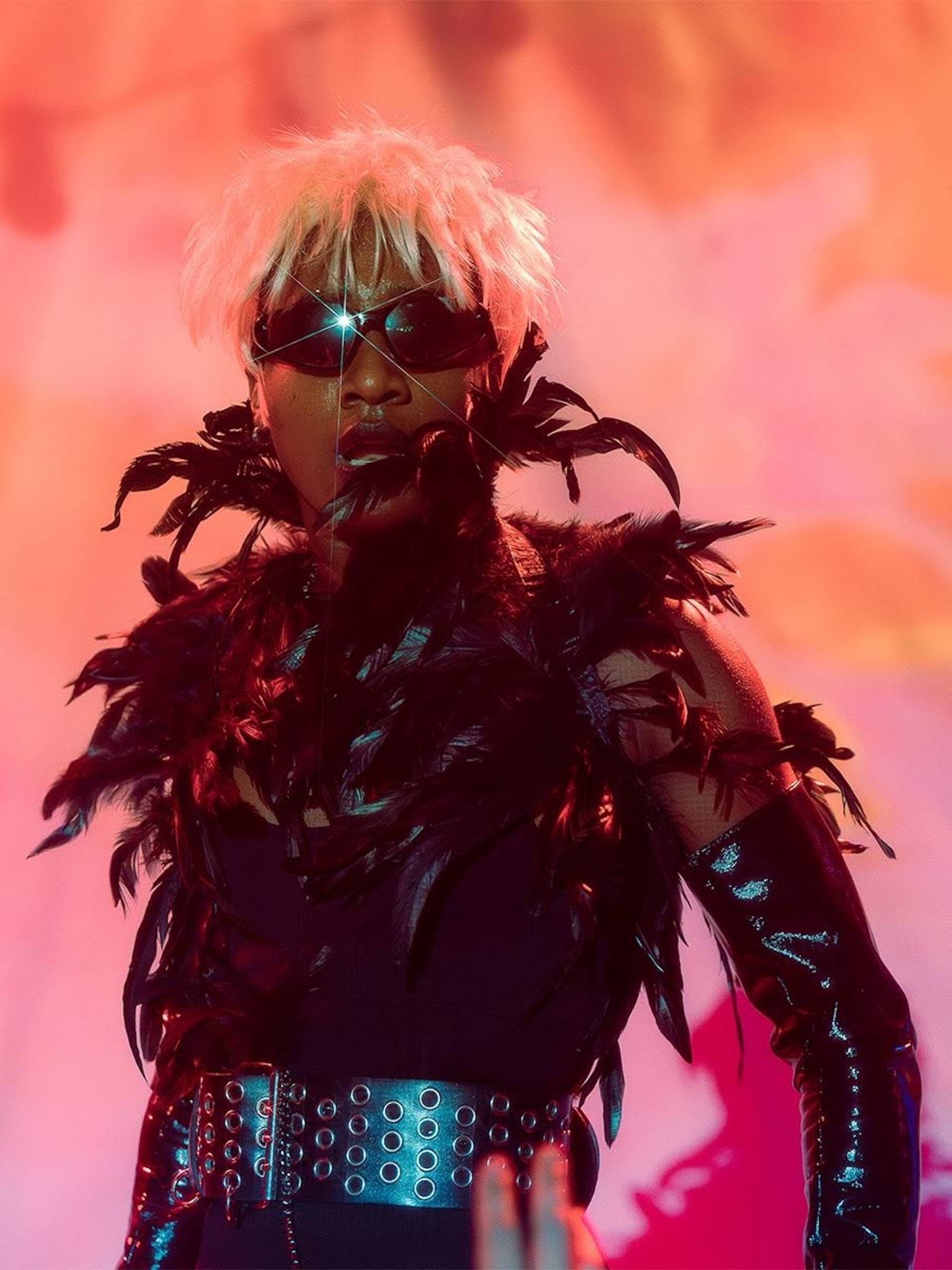A person in a black feathered top, big black belt and sunglasses poses in front of a flaming red background.