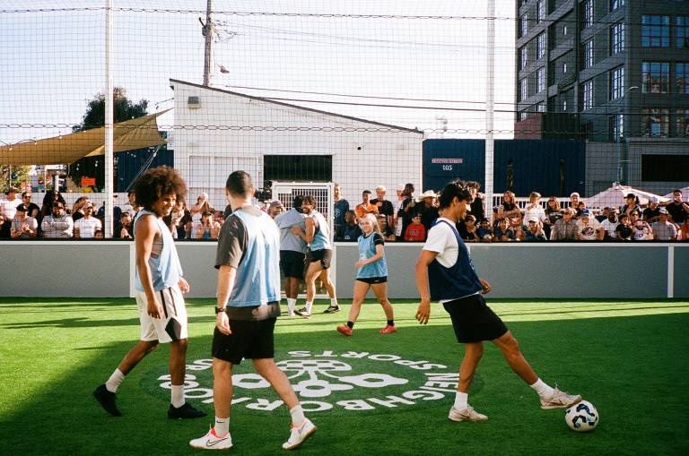 Six soccer players situate themselves on a green field, with spectators, netting and a slanted white building in the background