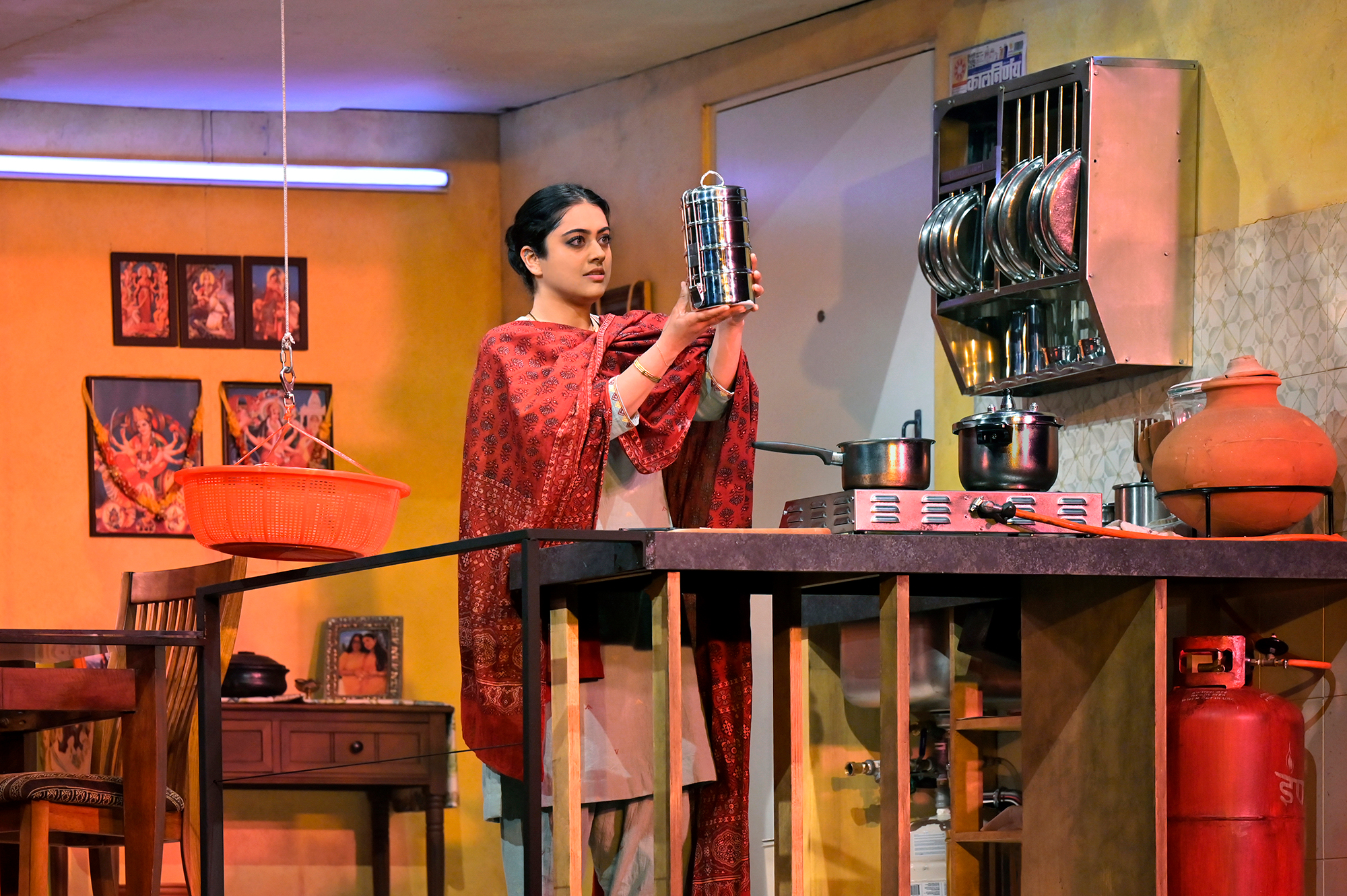 A young Indian woman holds a shiny silver cylinder aloft, wearing patterned fabric in a room decorated as a kitchen