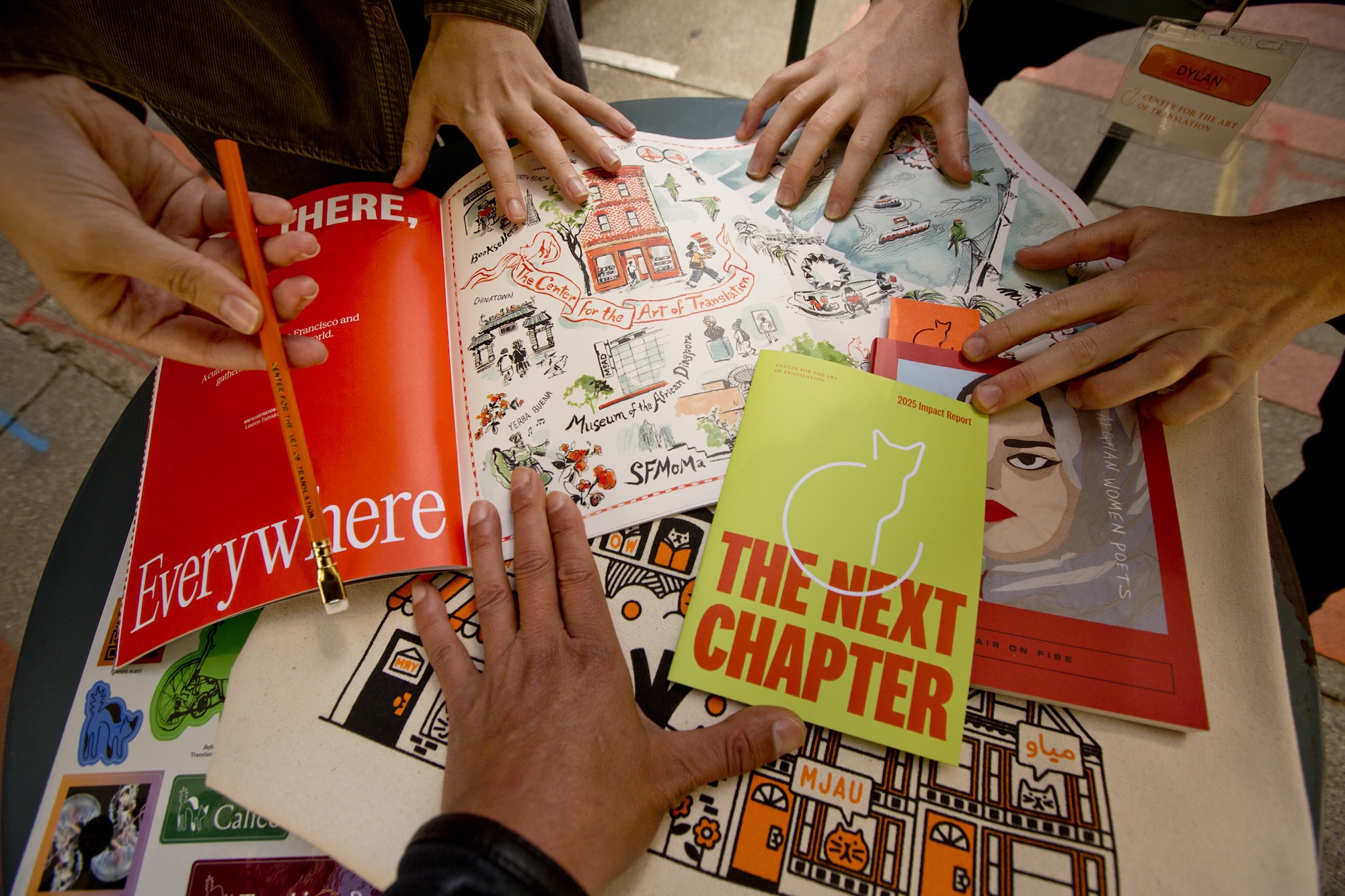 Three sets of hands on multiple illustrated publications, spread out on a table. One hand is holding a pencil.
