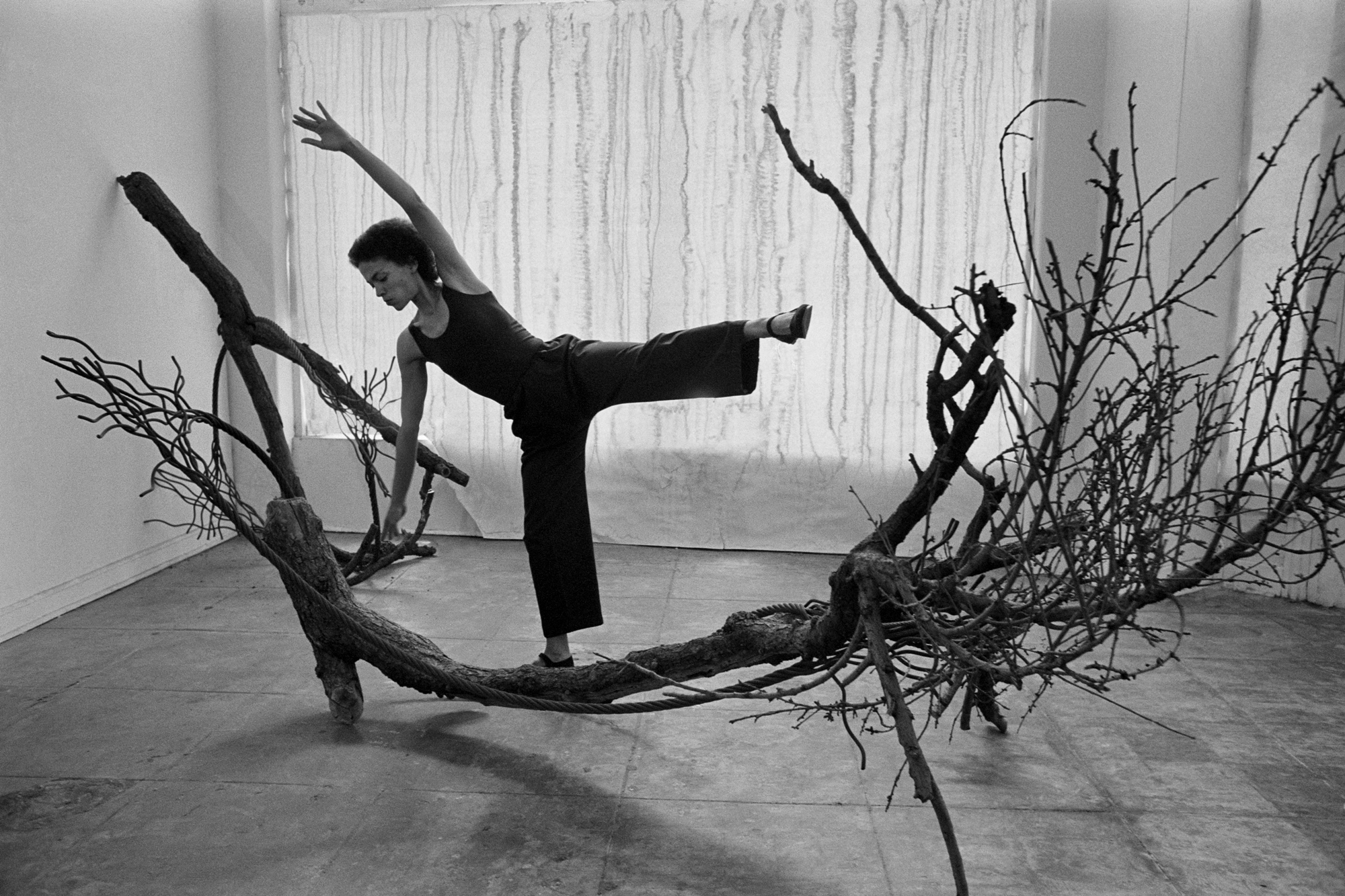 black-and-white image of Black woman posing with leg and arm raised to mimic large tree branch she stands on