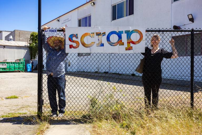 two people stand behind chainlink fence with "scrap" sign
