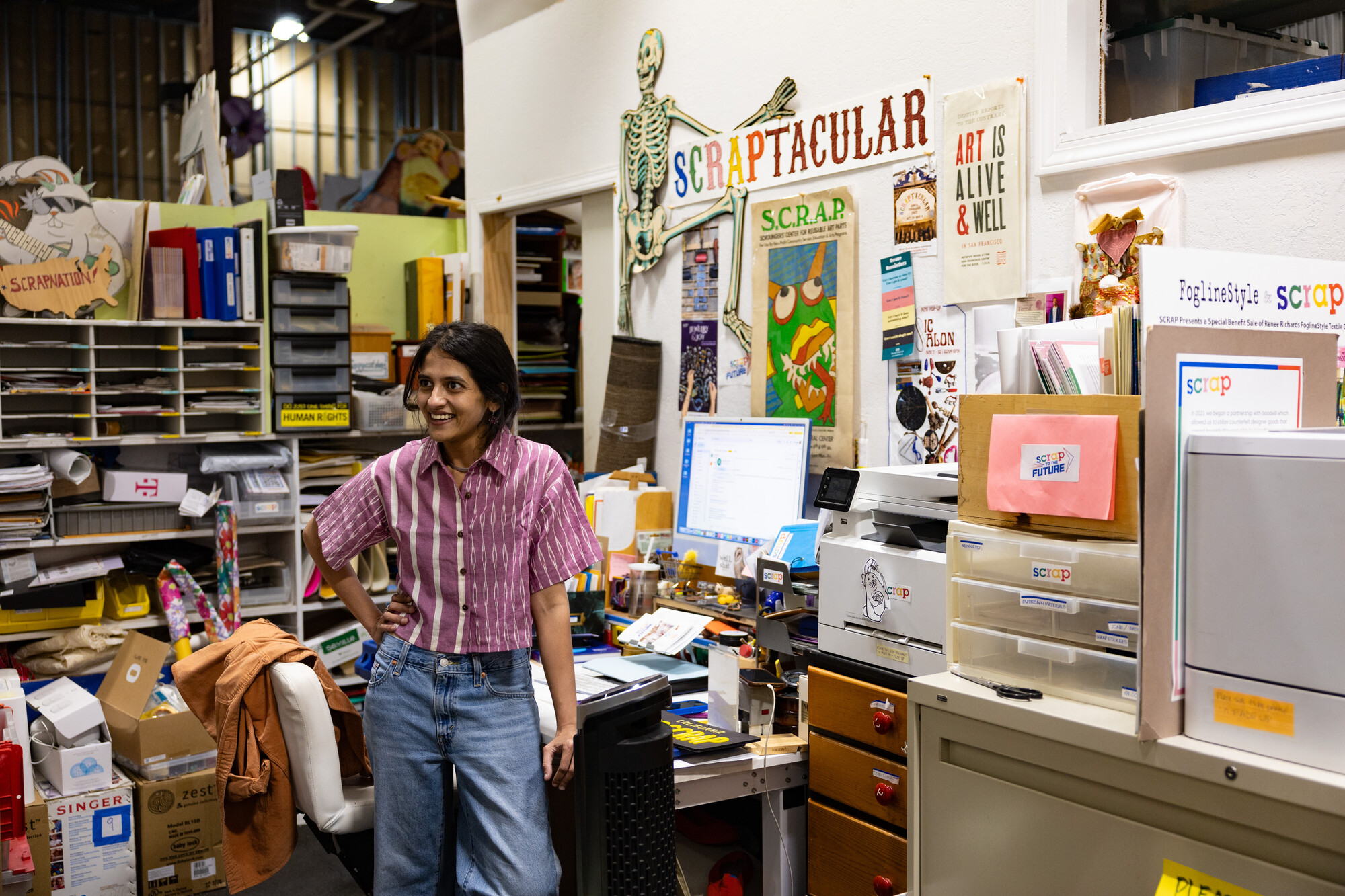 South Asian woman stands in office filled with boxes and posters