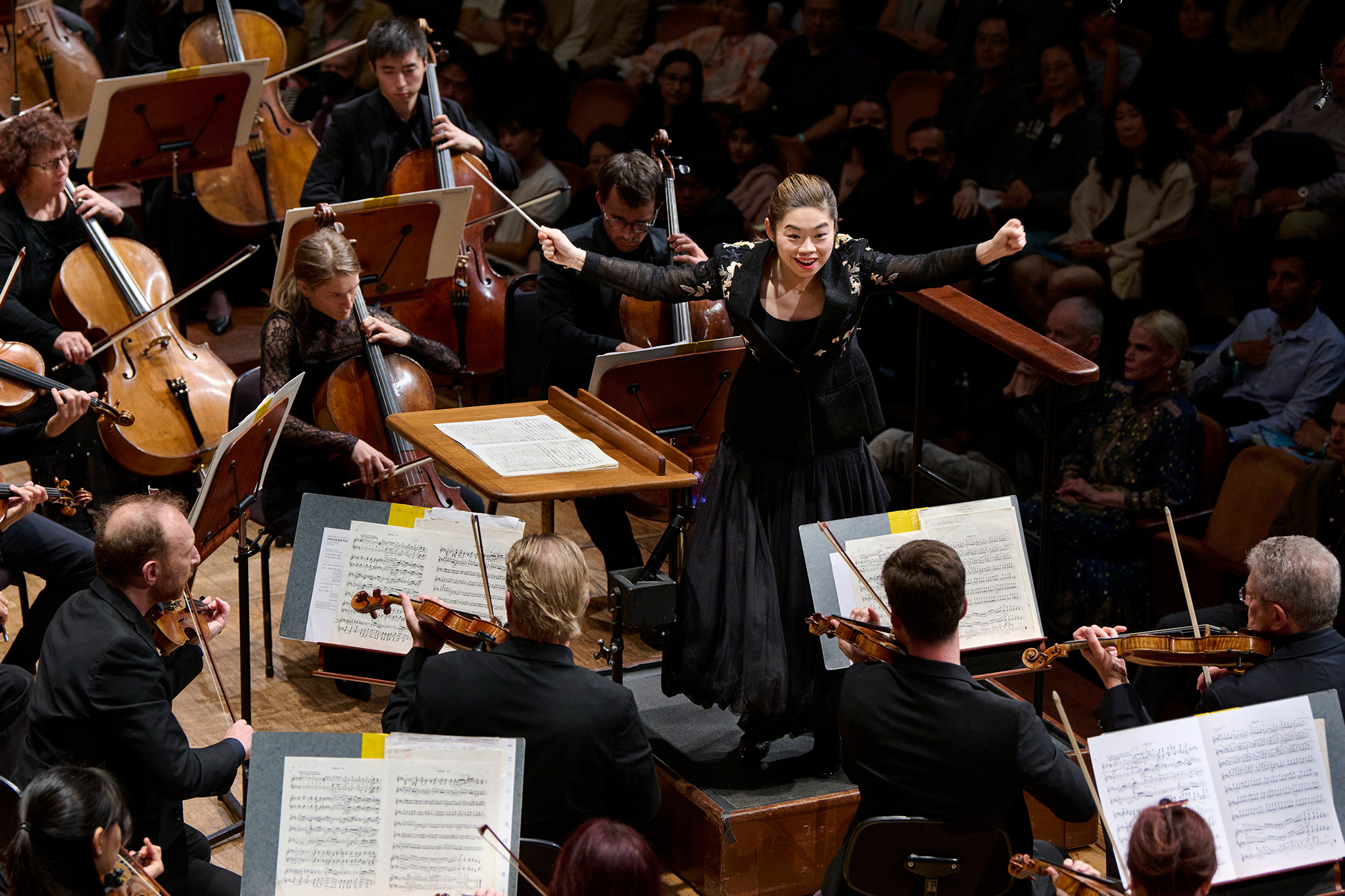 A young Asian woman stands, arms outstretched, holding a baton in front of a symphony orchestra
