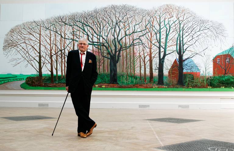 A man in a suit leans on a cane in front of a painting of trees