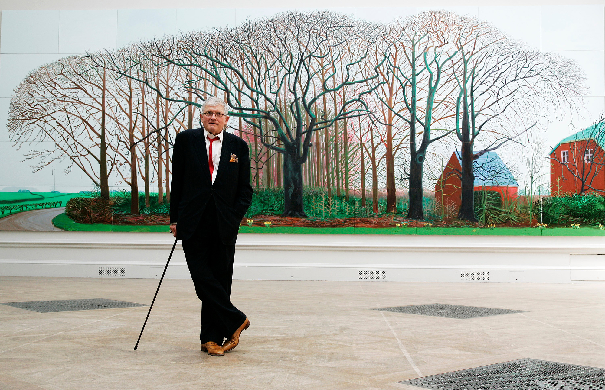 A man in a suit leans on a cane in front of a painting of trees