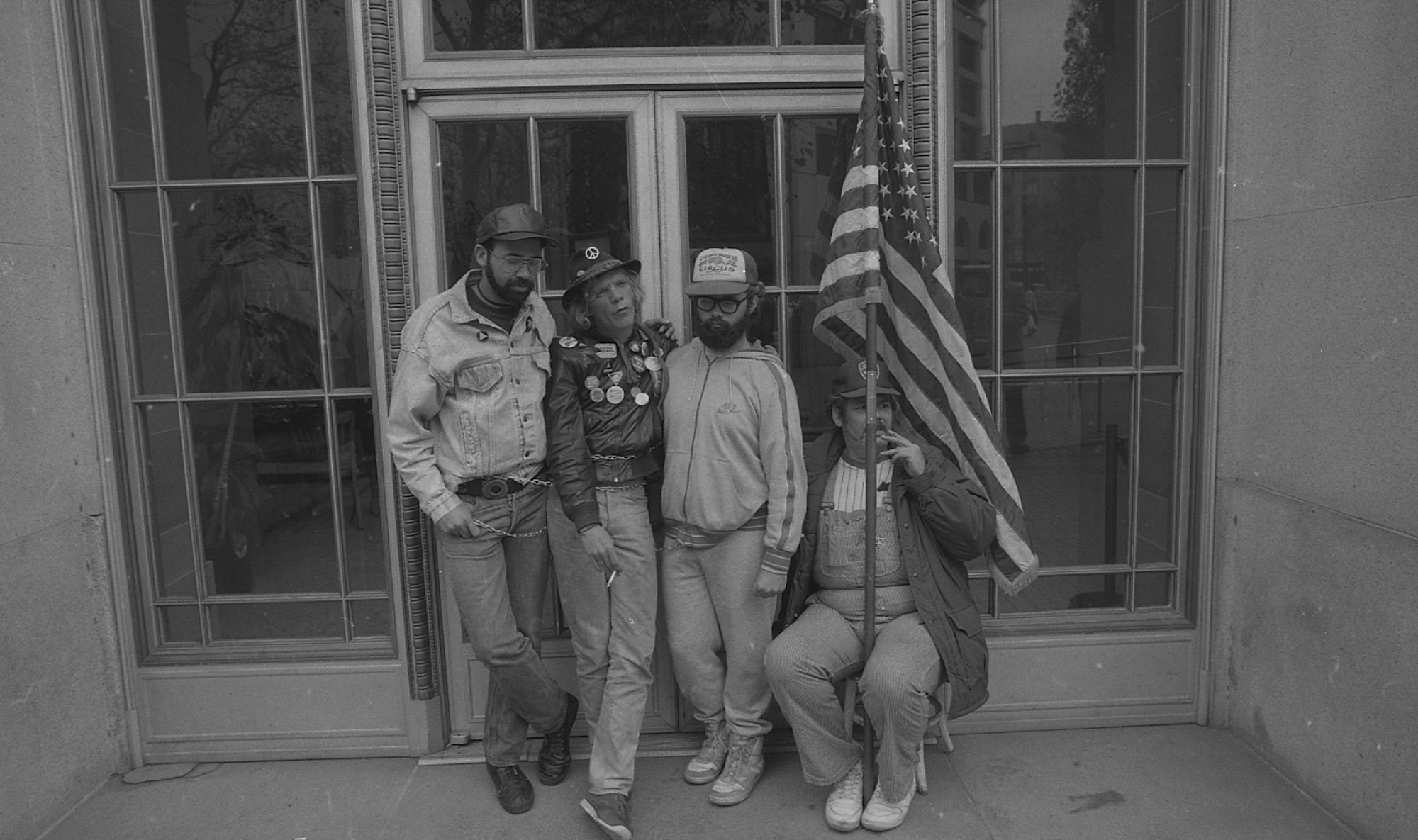 Four men of varying ages stand outside doors to an office building. One is holding a large American flag.