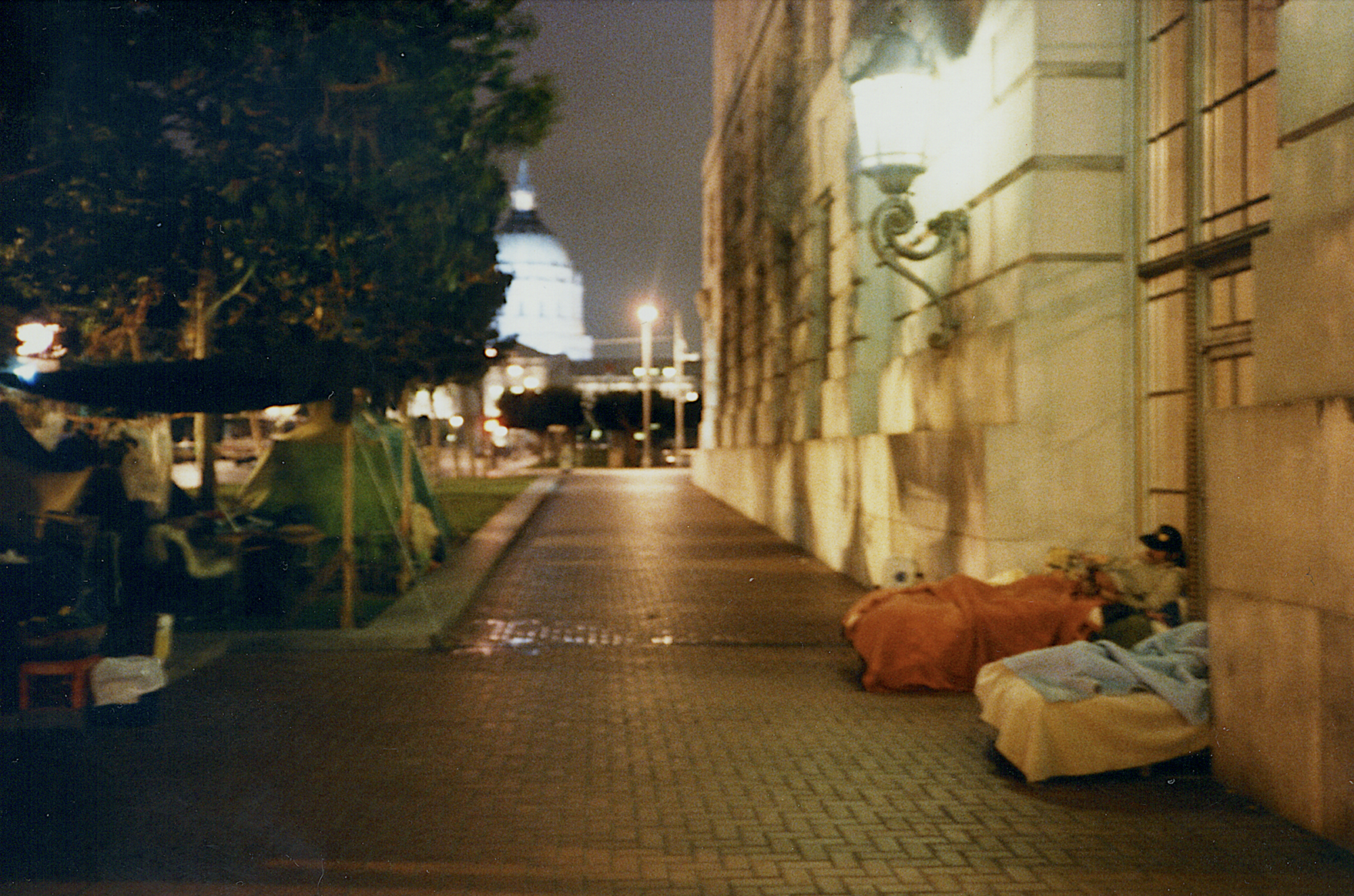 A government building with two mattresses visible in its doorway. Opposite, on a grassy area, several tents are set up.