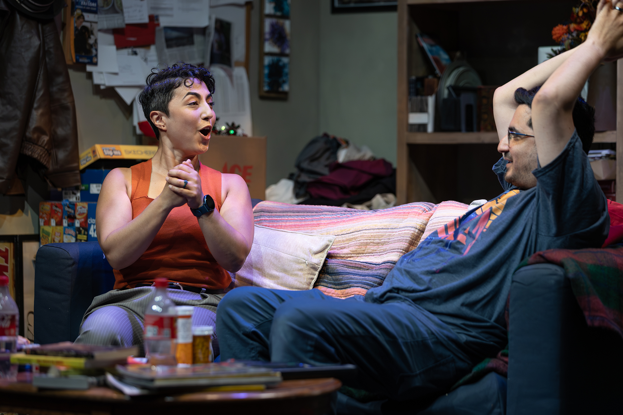 A woman and man sit on a couch, amid a very cluttered room, actively in conversation