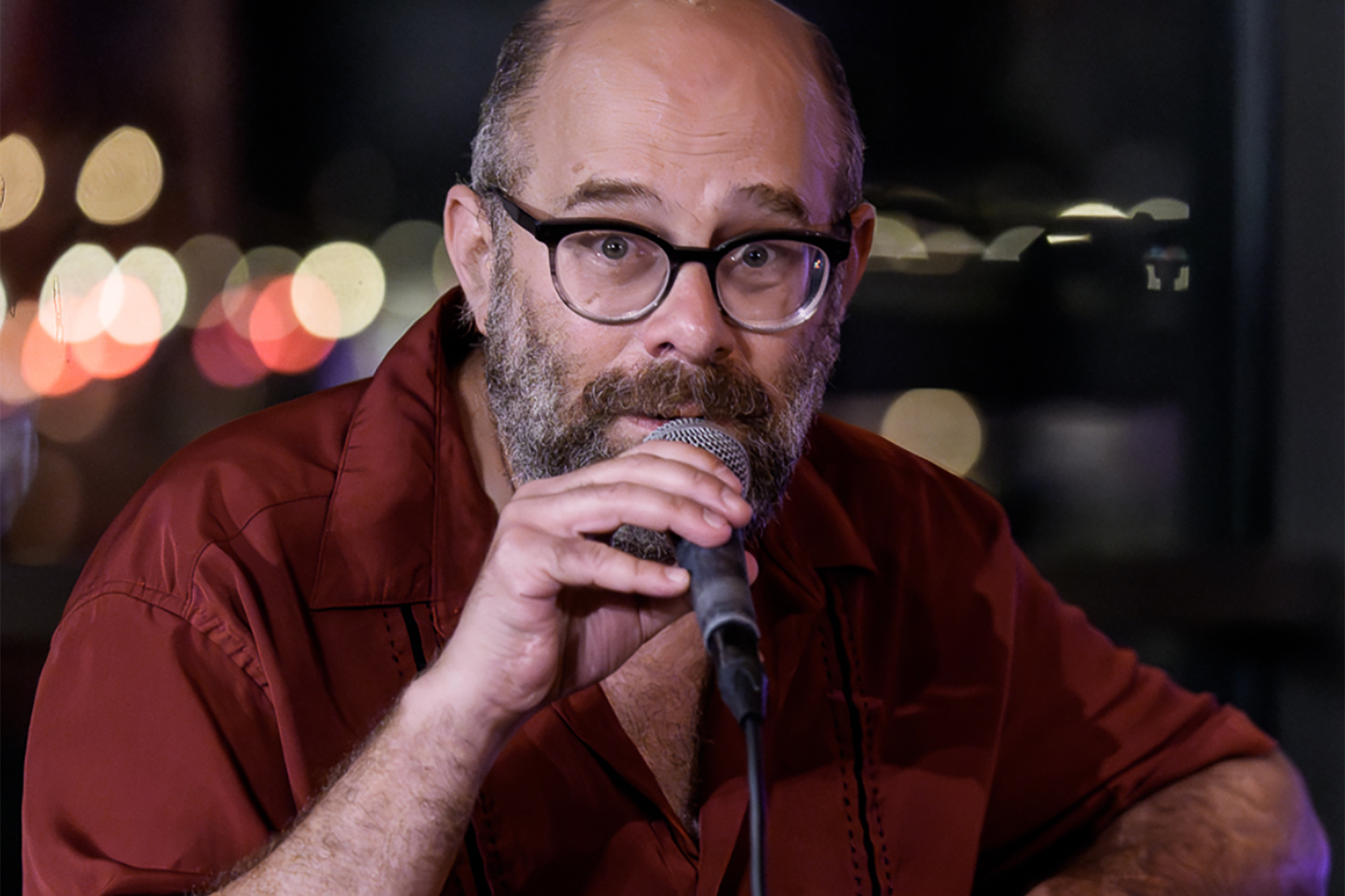 A man in a beard and deep burnt red shirt holds a microphone, looking ahead and singing