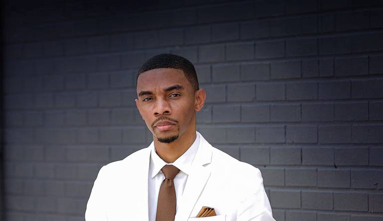 An African-American man in a white blazer and brown tie poses for a photo.
