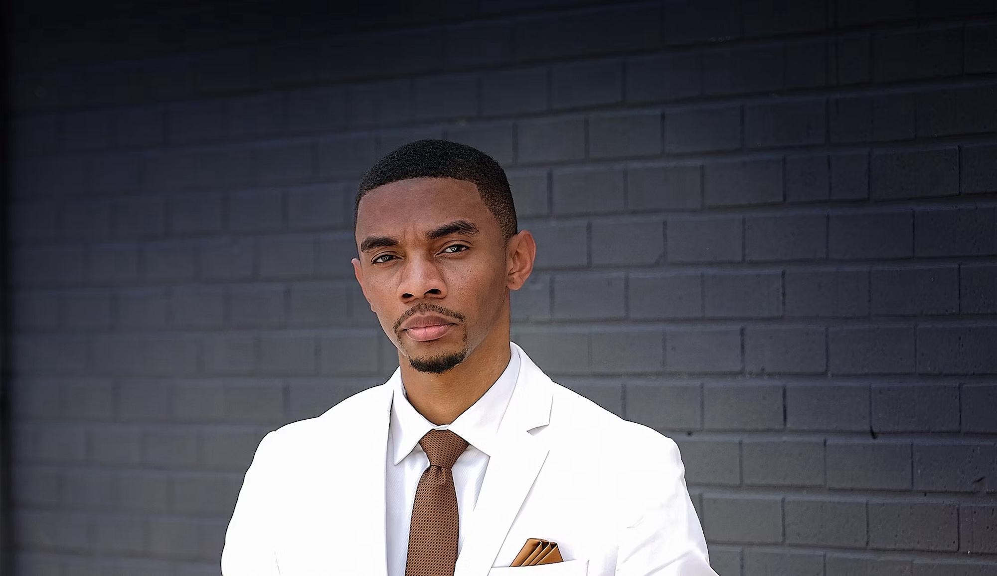 An African-American man in a white blazer and brown tie poses for a photo.
