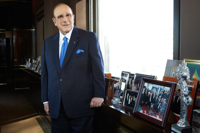A white man in a blue suit and tie stands in an office next to a table full of framed photos.