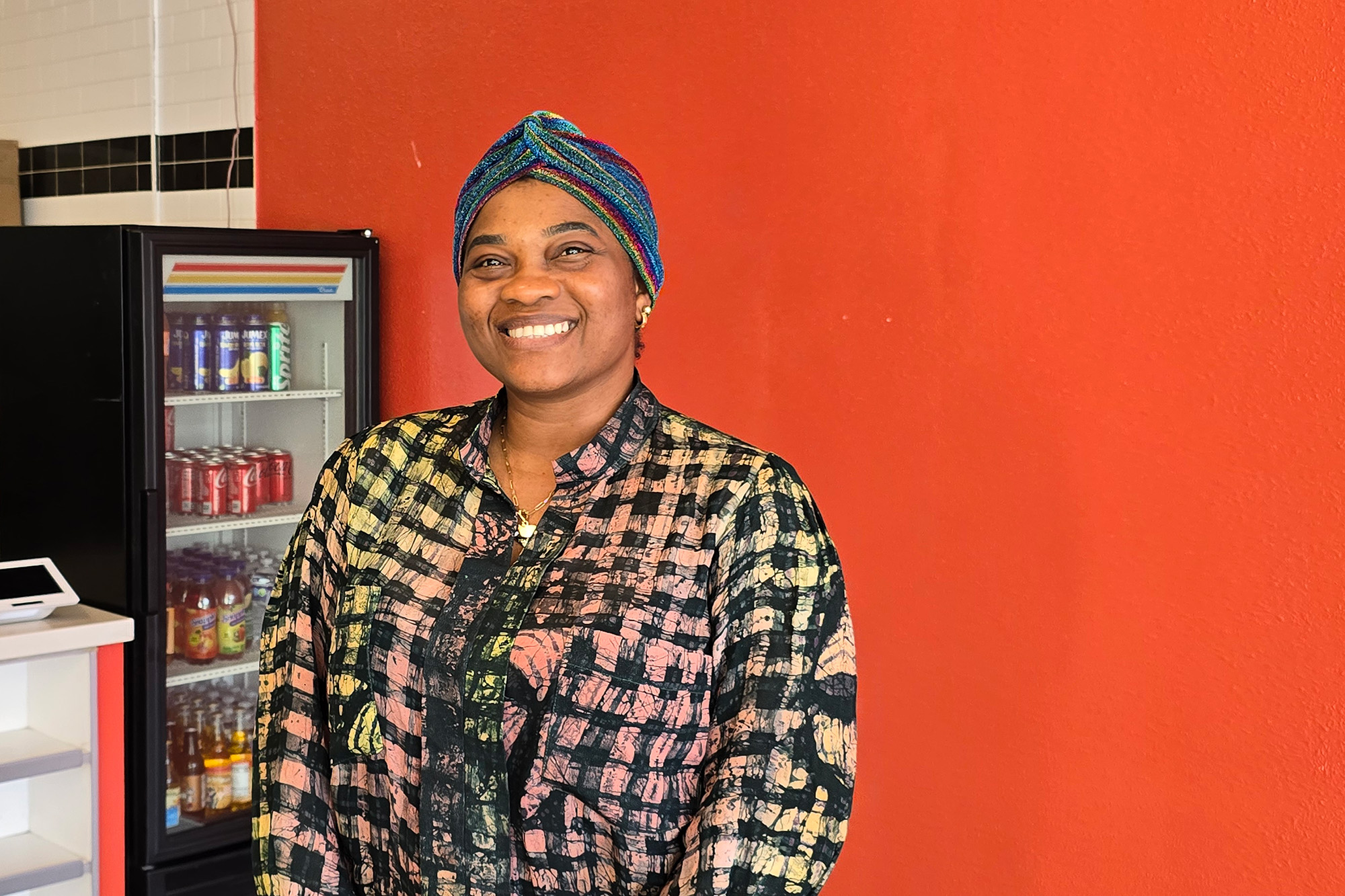A smiling Nigerian woman poses in front of an orange wall inside her restaurant.