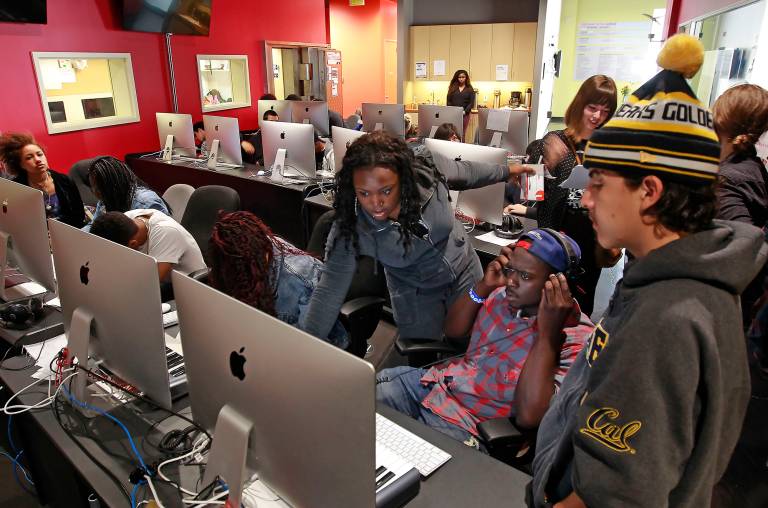 A young Black woman in blue denim helps a multiethnic group of students in front of a room of desktop computers.