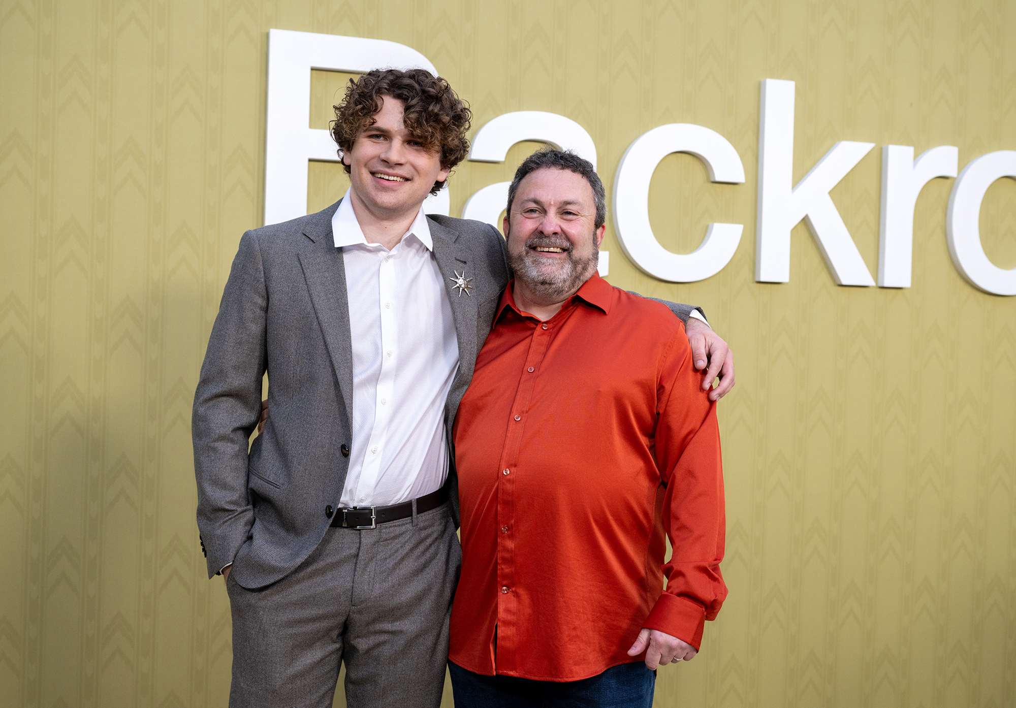 A young 20-year-old man in a suit and an older man in a red shirt pose in front of a drab yellow wall.