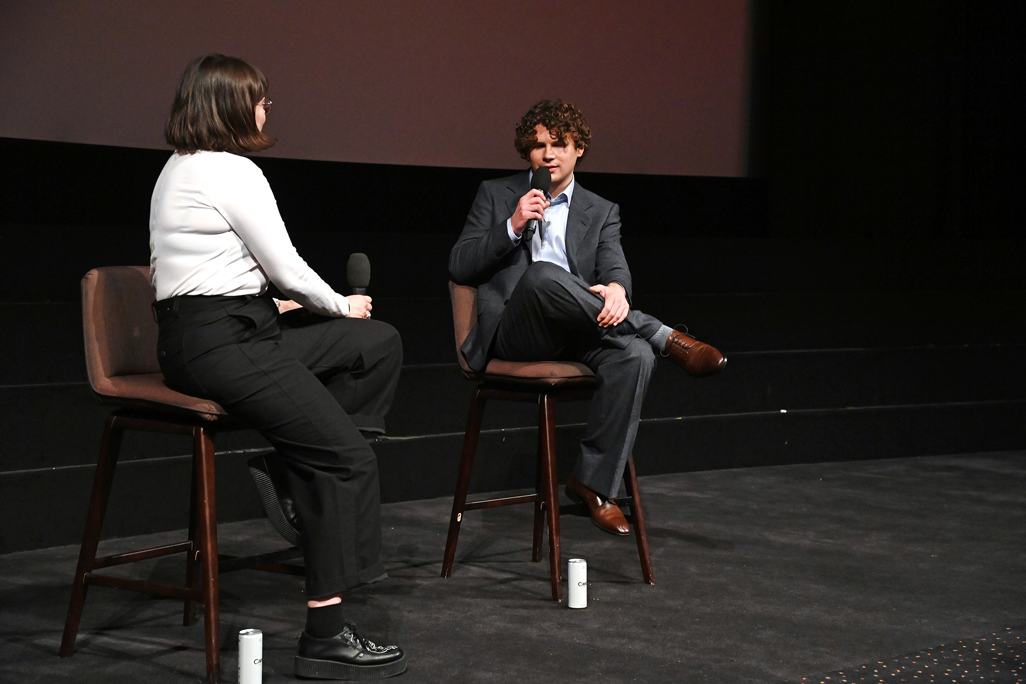 A young man in a suit sits holding a microphone, talking with a nearby woman, both in folding chairs