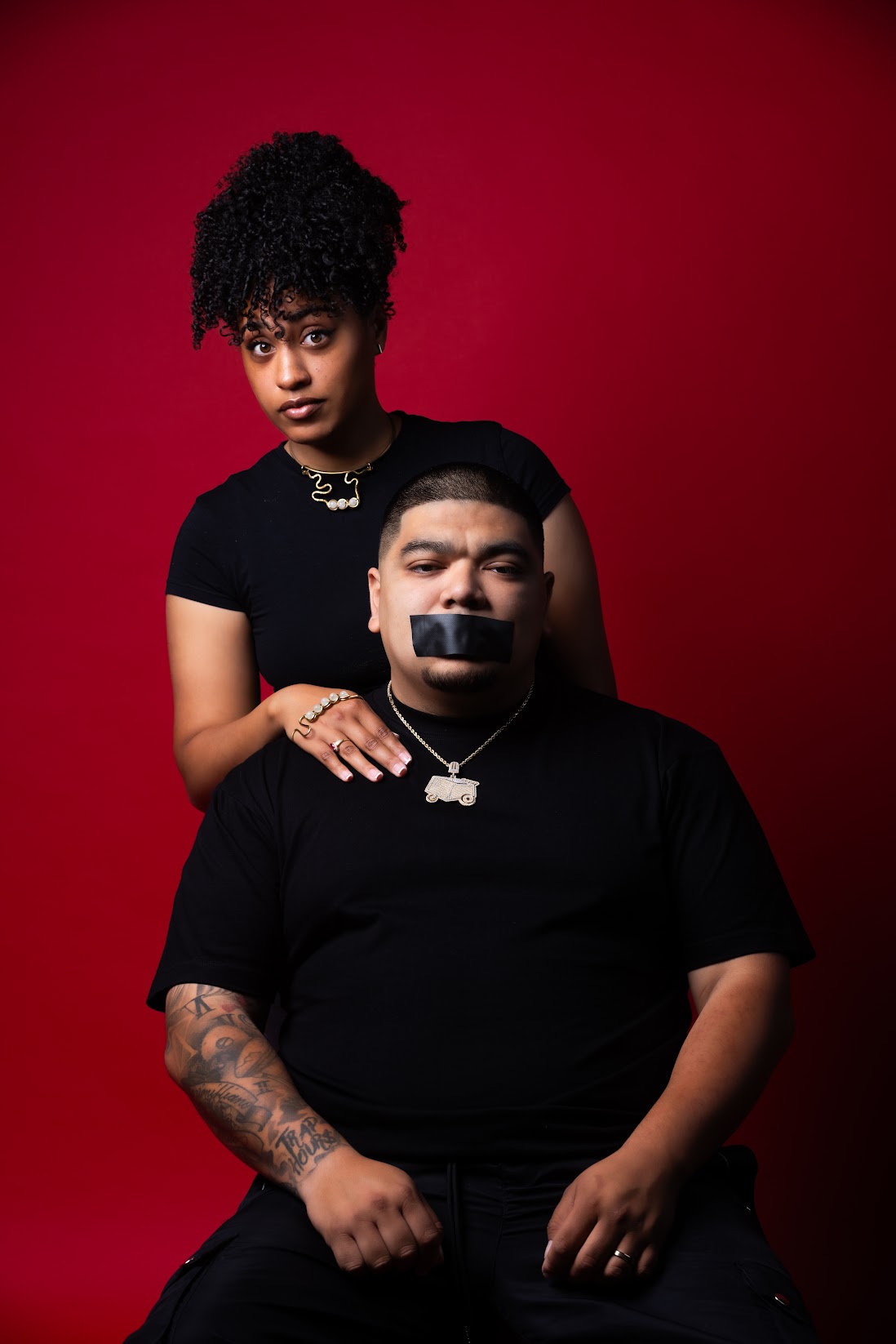 Two people, a man and a woman, pose for a photo. The man sits with a piece of black tape on his mouth while the woman stands behind him.
