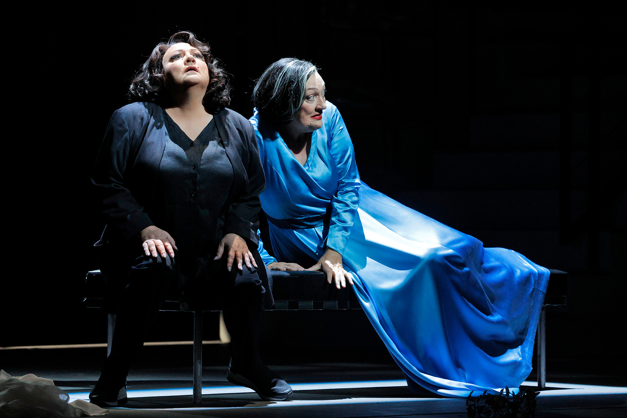 Two women in black and blue wardrobe sit on a bench, looking afraid