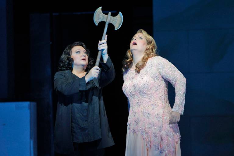 A woman in a black suit outfit hoists a double-edged axe in the air as a second woman in a light pink dress gazes up at it in a stupor