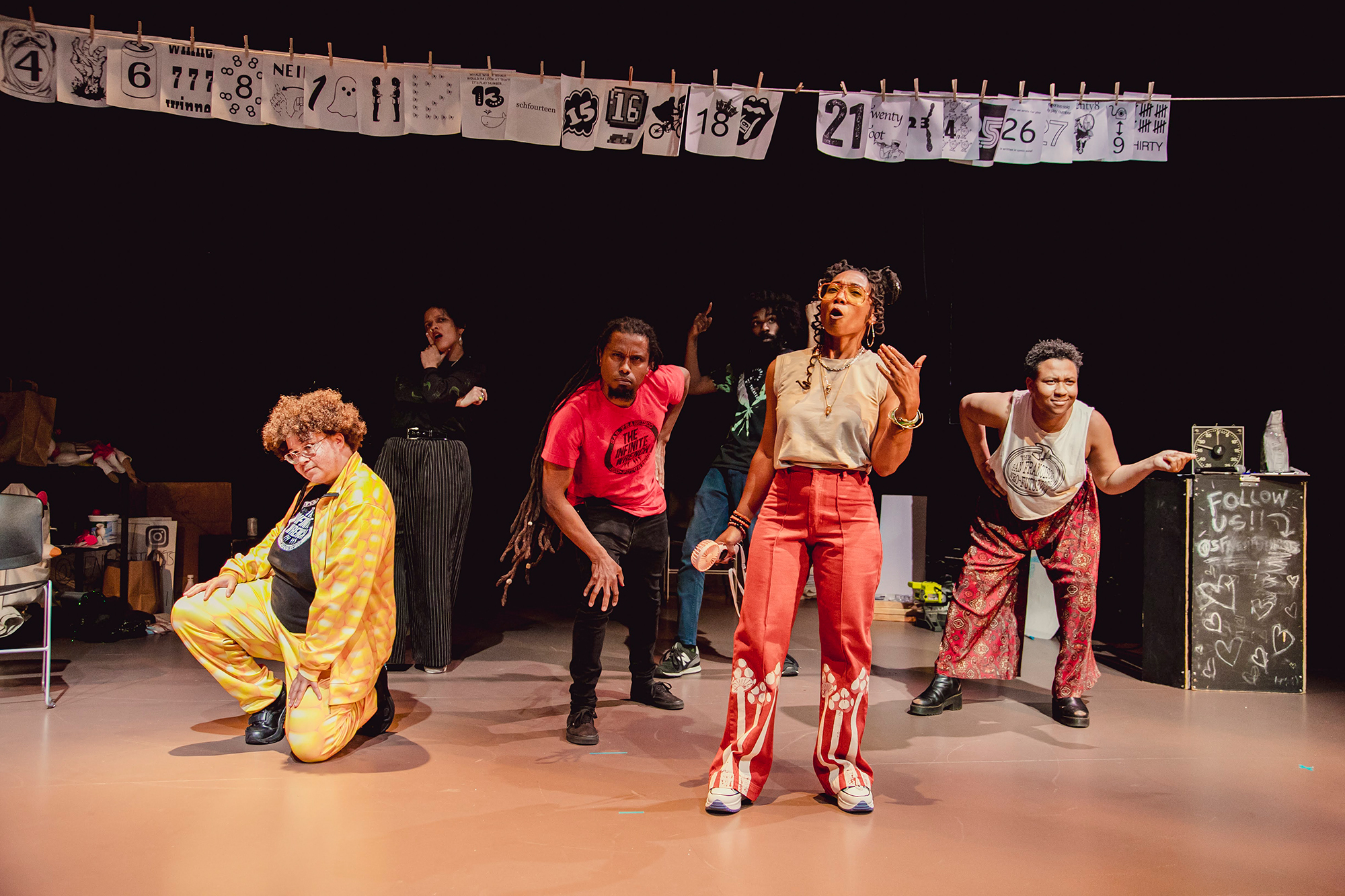 A group of Black actors on a beige wooden stage with a black background