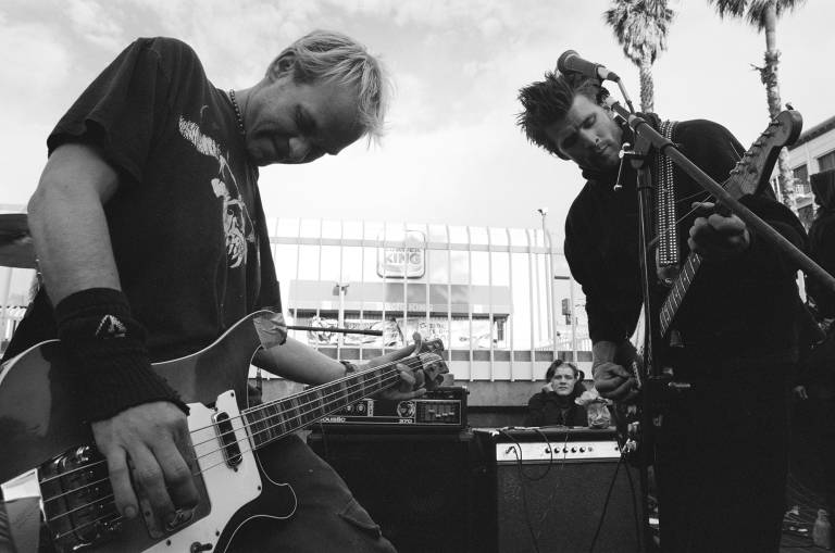 Two young men play electric bass and guitar against a backdrop of iron fencing and a Burger King