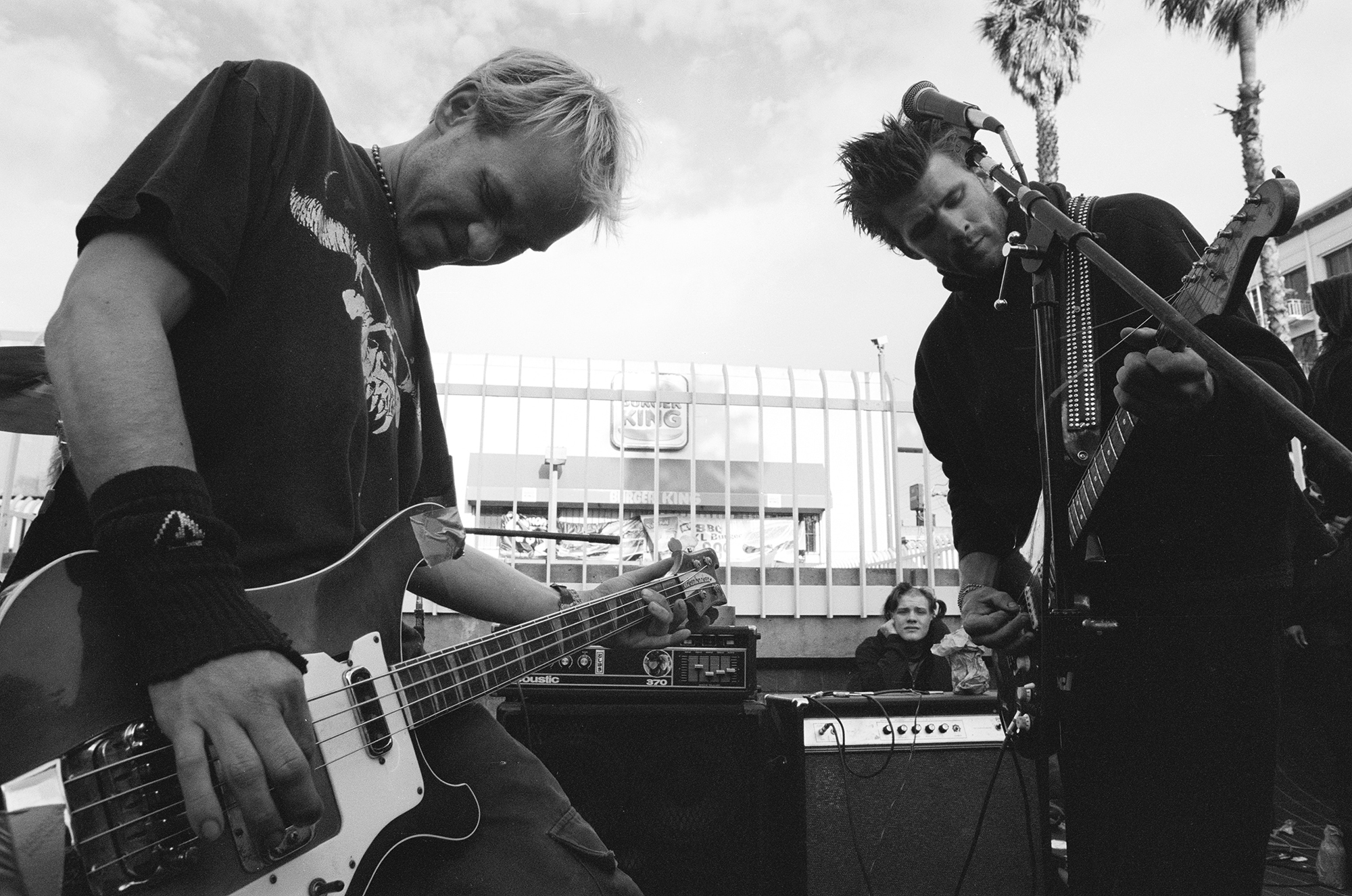 Two young men play electric bass and guitar against a backdrop of iron fencing and a Burger King