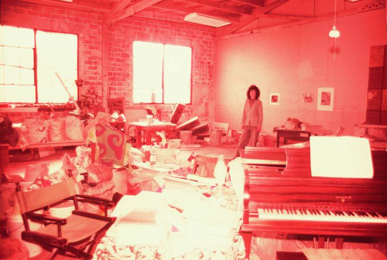 old snapshot of smiling woman in large material-filled studio space, piano in foreground