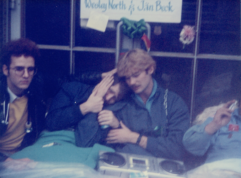 A grainy photograph of two men under a blanket, sitting up. One leans his head on the other. Two friends sit either side of the men.