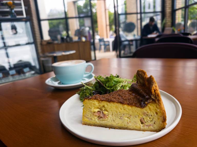 Thick slice of quiche and a cup of coffee on a table inside a sunny cafe.