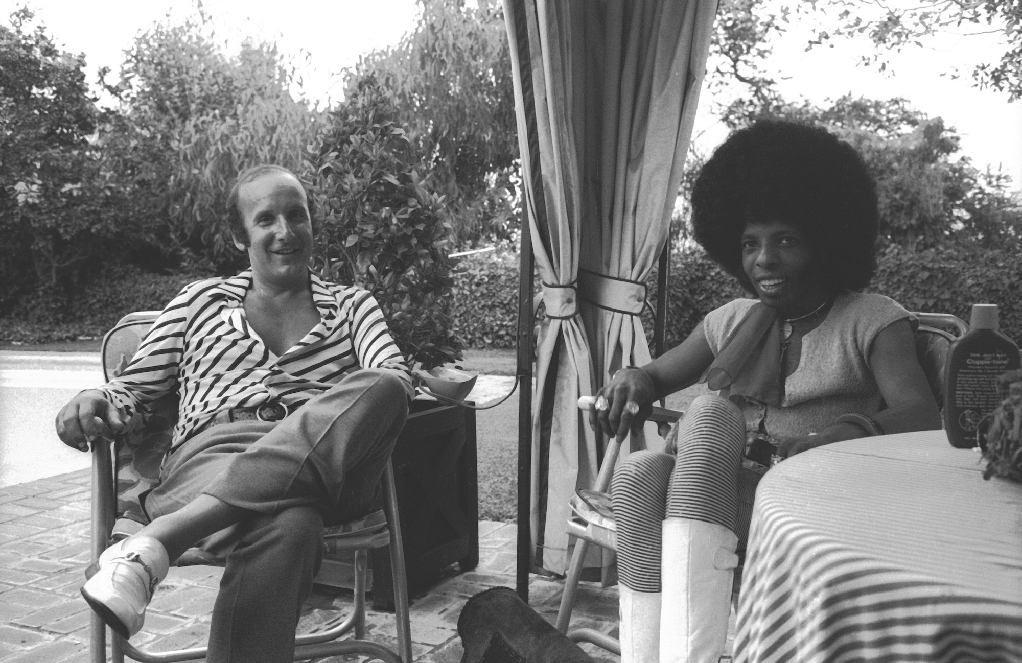 A white man wearing 1970s attire sits next to a Black man with a large afro and neck scarf. They are sitting outside on lawn chairs.