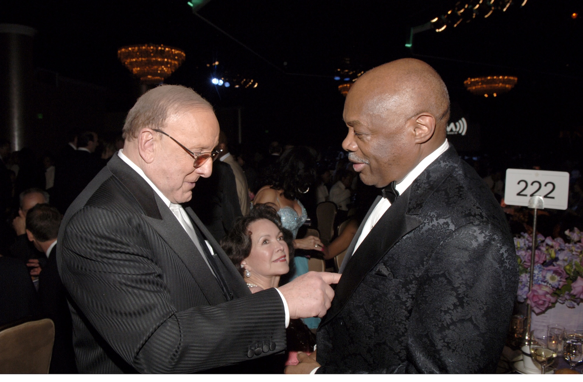 In a ballroom hosting a large dinner, a white man in a black suit points to the white shirt of a Black man, also wearing black tie.