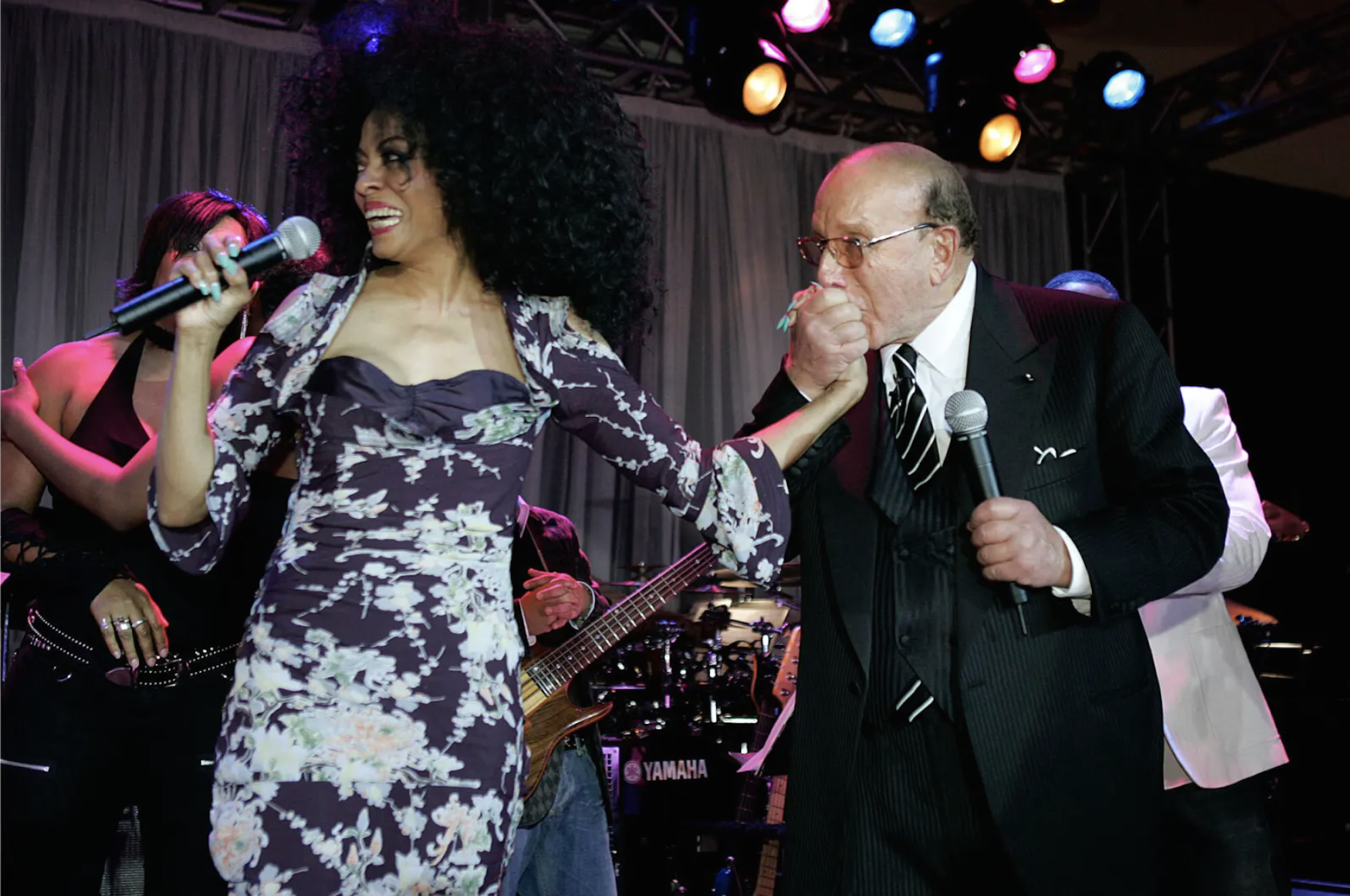 A very slender Black woman wearing a skintight black and floral dress stands on stage smiling and holding a microphone, as a white man in a black suit warmly kisses her hand. He is also holding a mic.