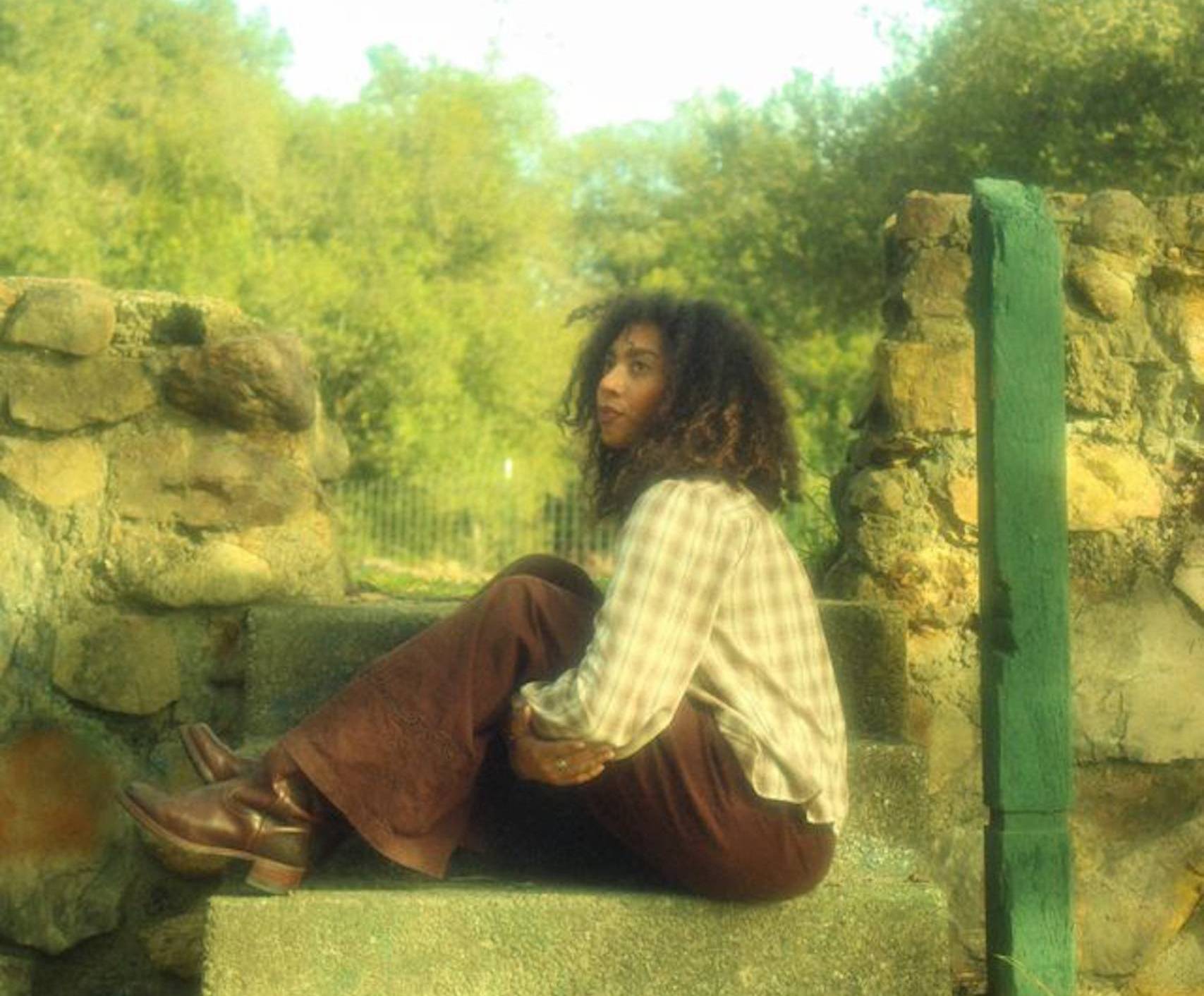 A woman sitting on a set of concrete stairs, wearing brown pants and a plaid shirt, poses for a photo.