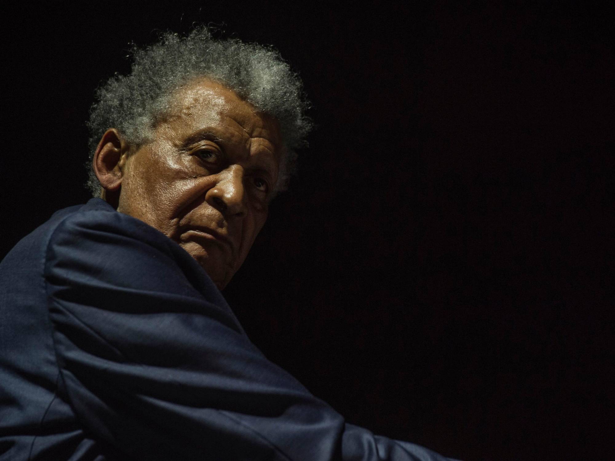 A Black man with greyish hair and a dark shirt looks over his right shoulder while at the piano, against a black background