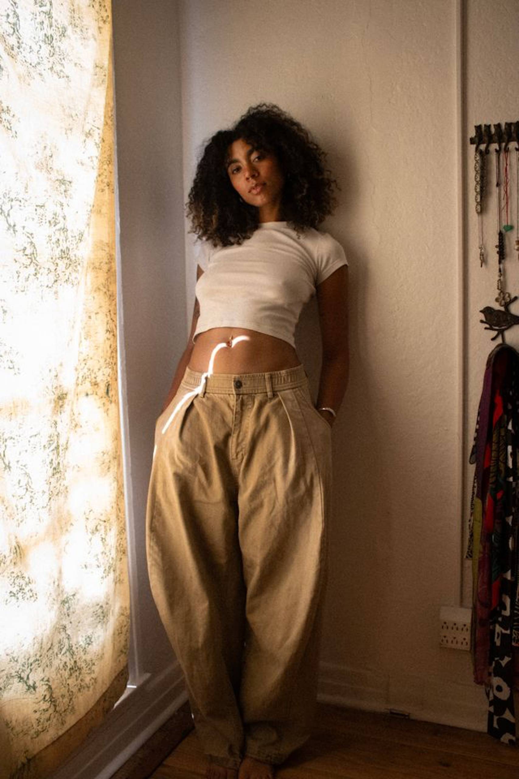 A woman stands inside of a house, against a wall and near a window, wearing a white shirt and khaki pants, as she looks into the camera.