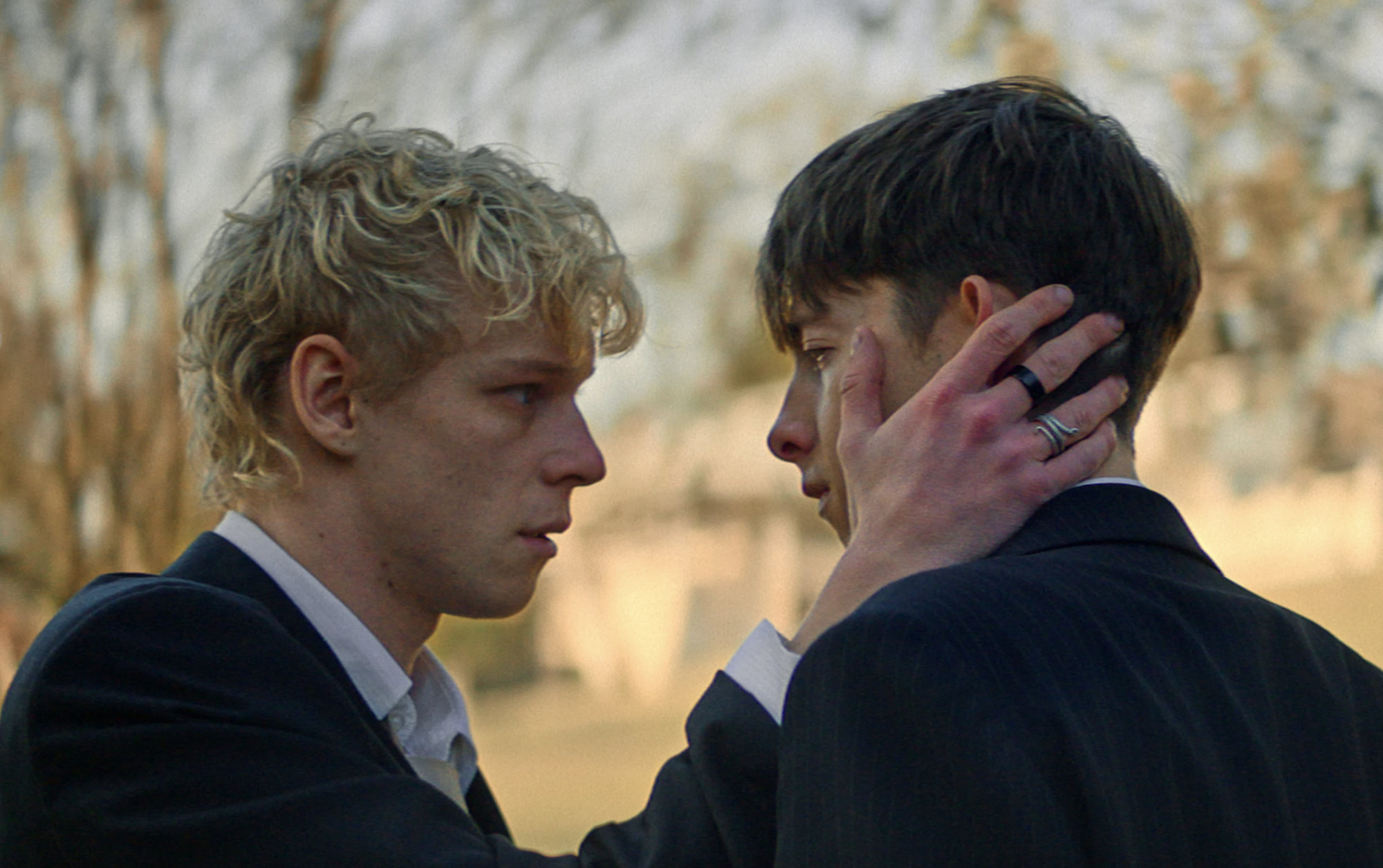 Two young white men look longingly at each other. One holds a cut hand up to the face of the other.