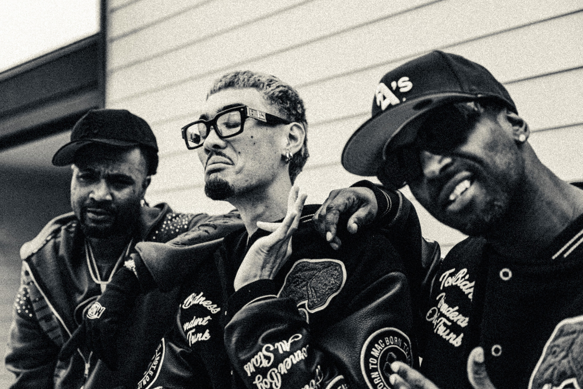 Black and white photo of three rappers in dark leather jackets, posing in front of a house.