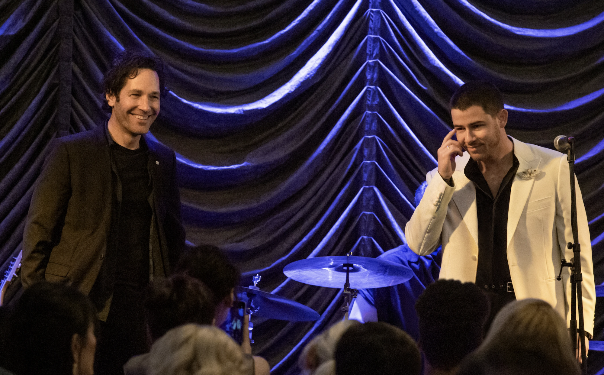 Two handsome white men stand side by side on stage together in front of a backlit purple curtain. They are both smiling.