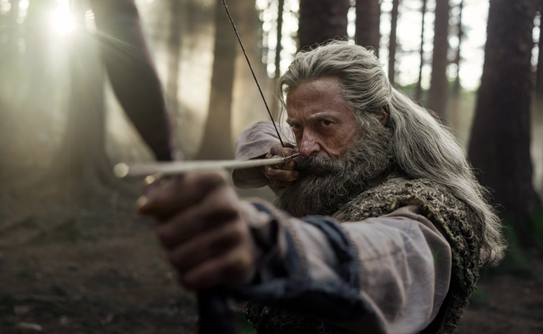 A grizzled white man with long grey hair stands in the woods with a bow and arrow aimed at something in the distance.