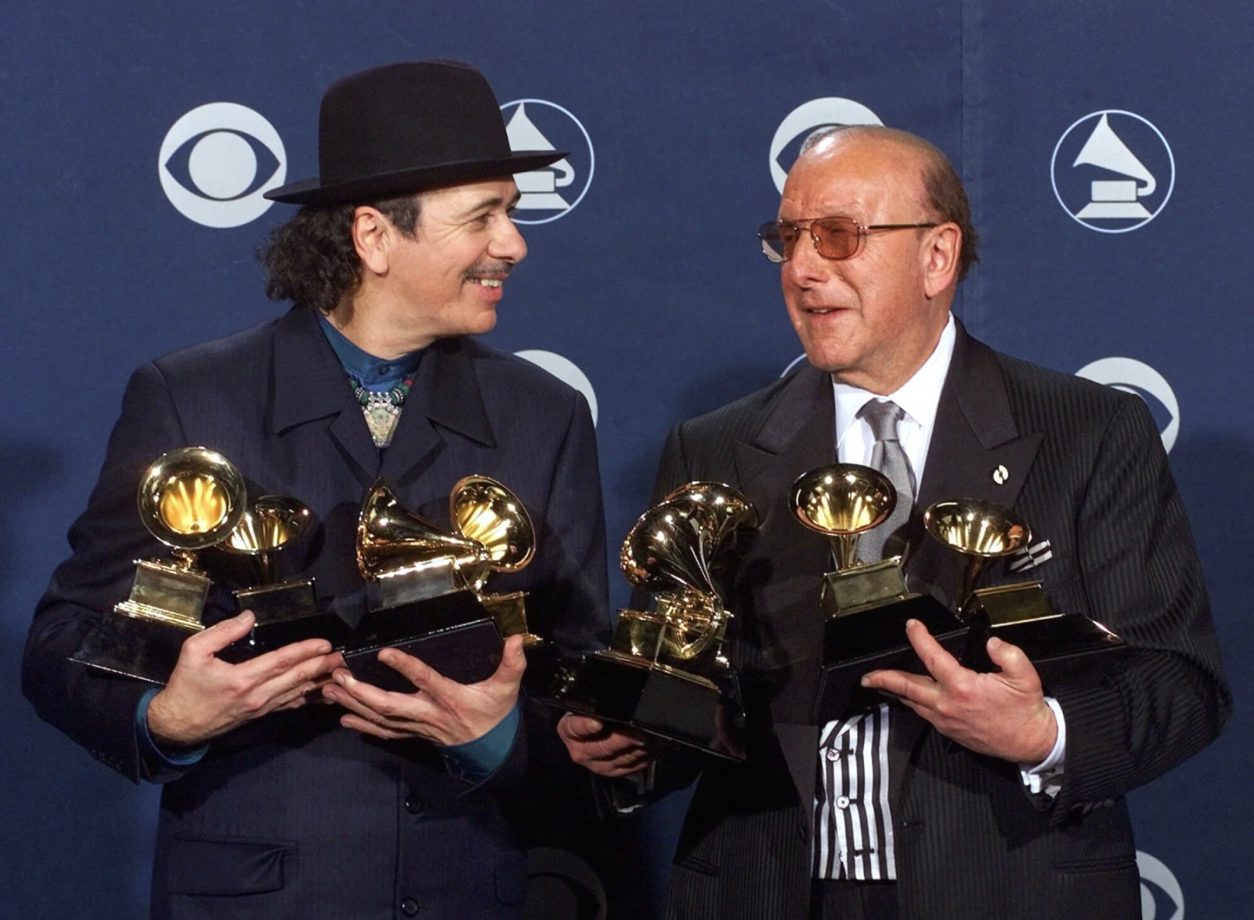 A Latino man wearing a hat and dark blue suit clutches four Grammy awards while looking to his left at a bald white man in a black suit. He is holding 3 Grammy awards.