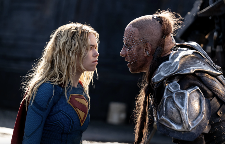 A young blond woman wearing a Supergirl uniform stands face to face with a man who resembles a cyborg, studs all over his face.