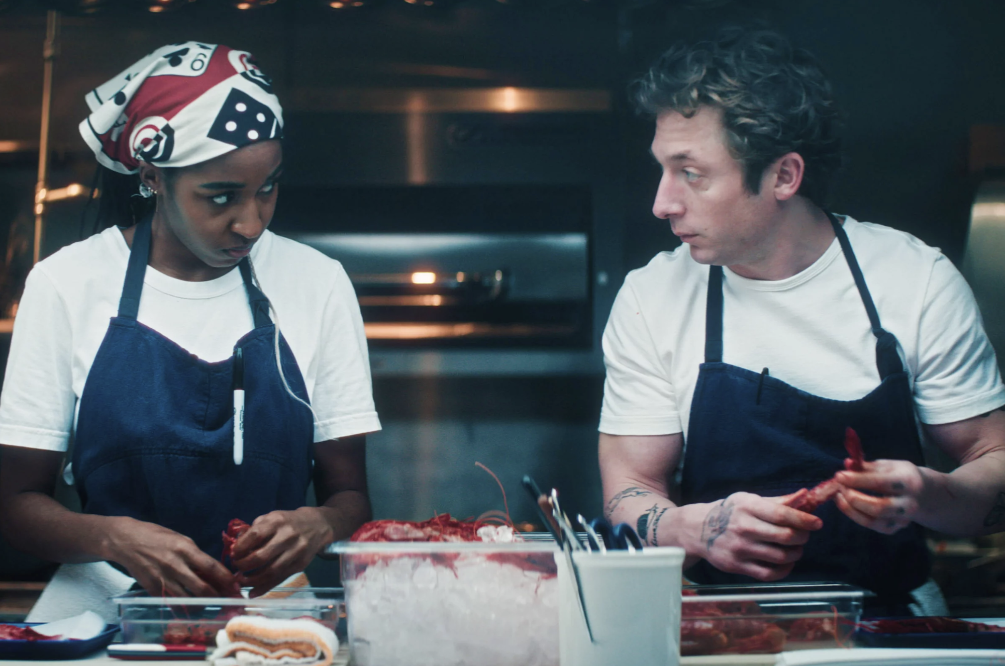 A Black woman wearing a headscarf, white t-shirt and blue apron looks at a white man, also wearing a white t-shirt and apron. They are standing side by side at a counter in a professional kitchen, shelling shrimp.