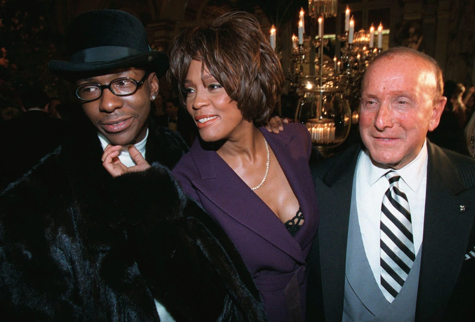 A Black man wearing a black bowler hat and fake fur coat smiles as a Black woman wearing a low cut purple suit jacket touches his chin. Next to them is an older white man wearing a striped tie and formal suit.
